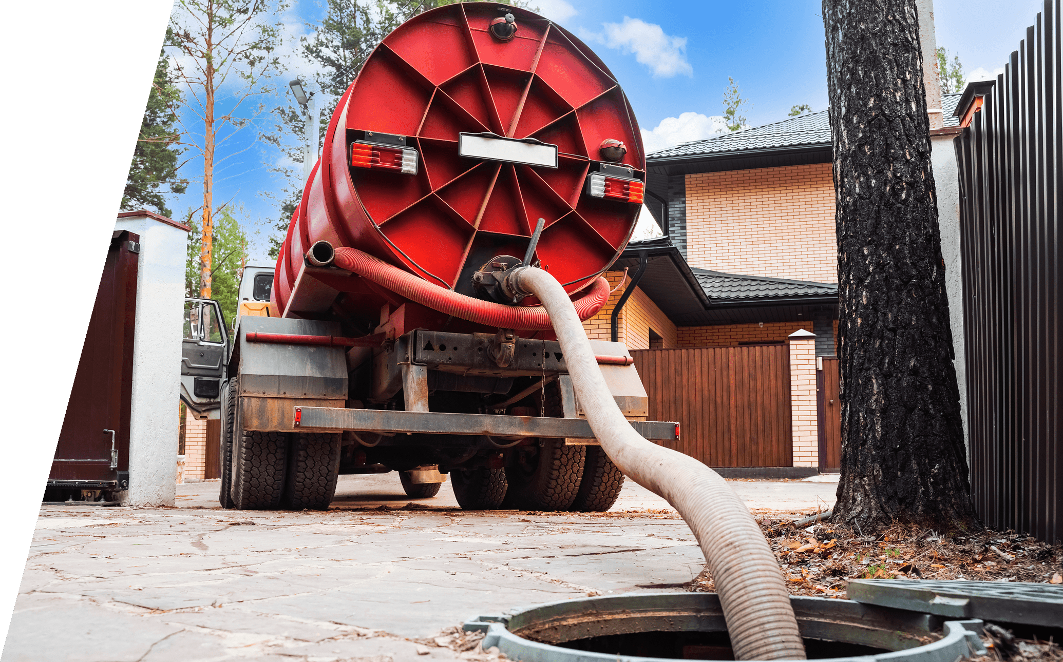 A septic tank truck with a red cylindrical container is parked, with a hose extending into an open sewer at a residential area.